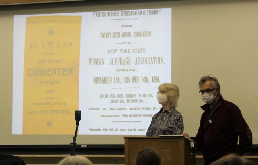 Elaine Engst and Laurent Saloff-Coste standing at a podium. The slide projected behind them displays an image of a New York State Woman Suffrage Association poster.